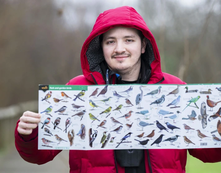 Smiling man in a red jacket holding a bird identification poster with various bird species displayed. Smiling man in a red jacket holding a bird identification poster with various bird species displayed.